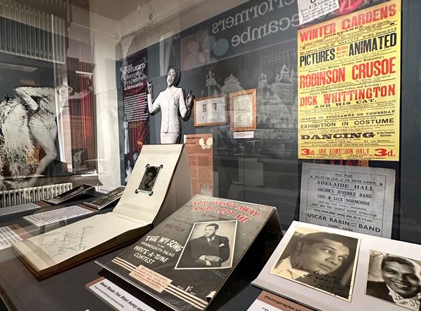 photo of exhibition room with life size figures of black performers on the walls and various objects in display cases. 